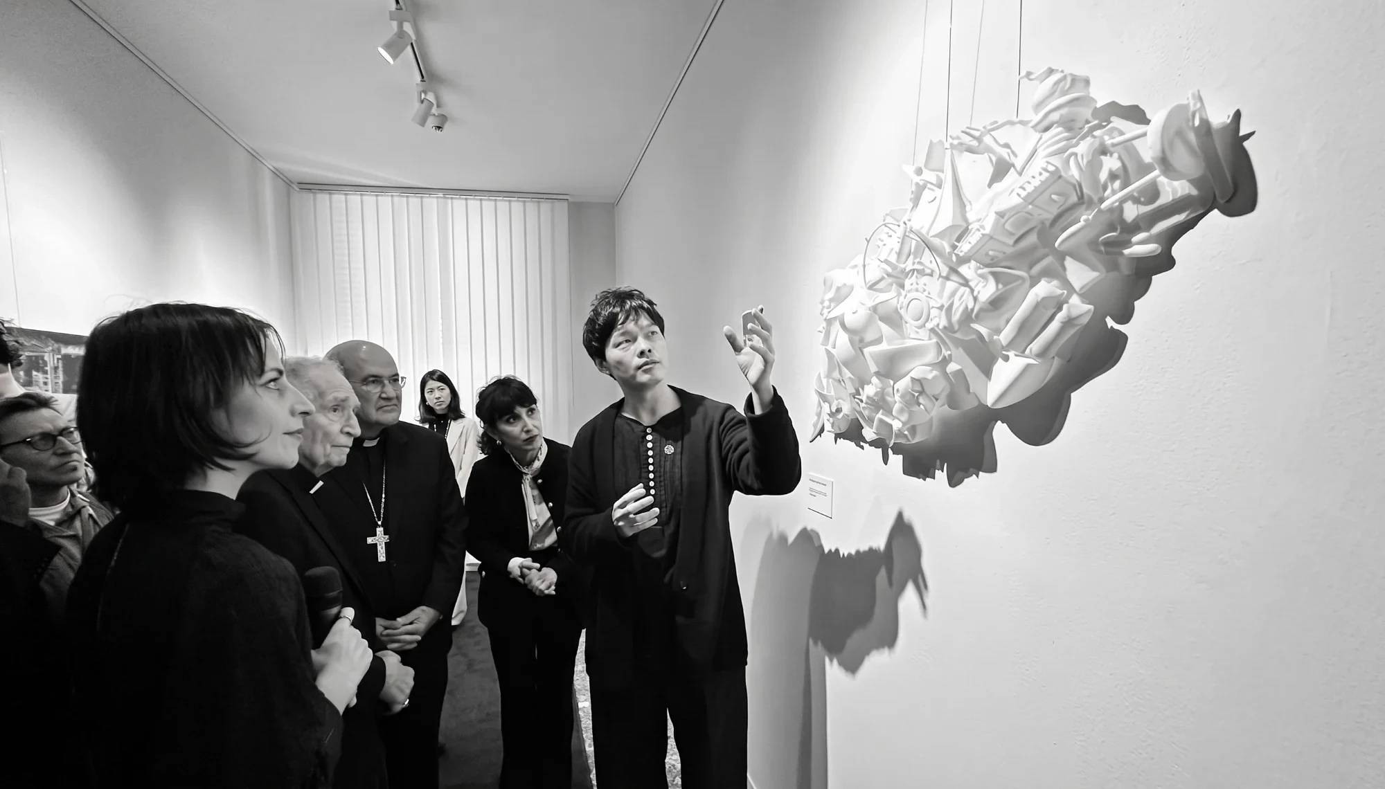 Black-and-white photo of Chien-hua Huang presenting his white sculpture to visitors at the Interconnected Hearts exhibition, 2024.