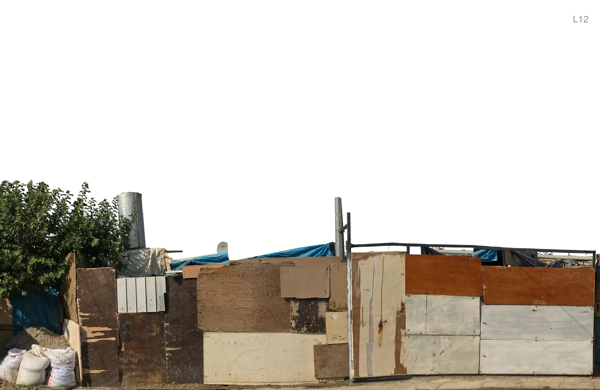 The street front of a camp home, patched together from plywood, metal sheets and blue tarpaulin, with sacks on the ground.