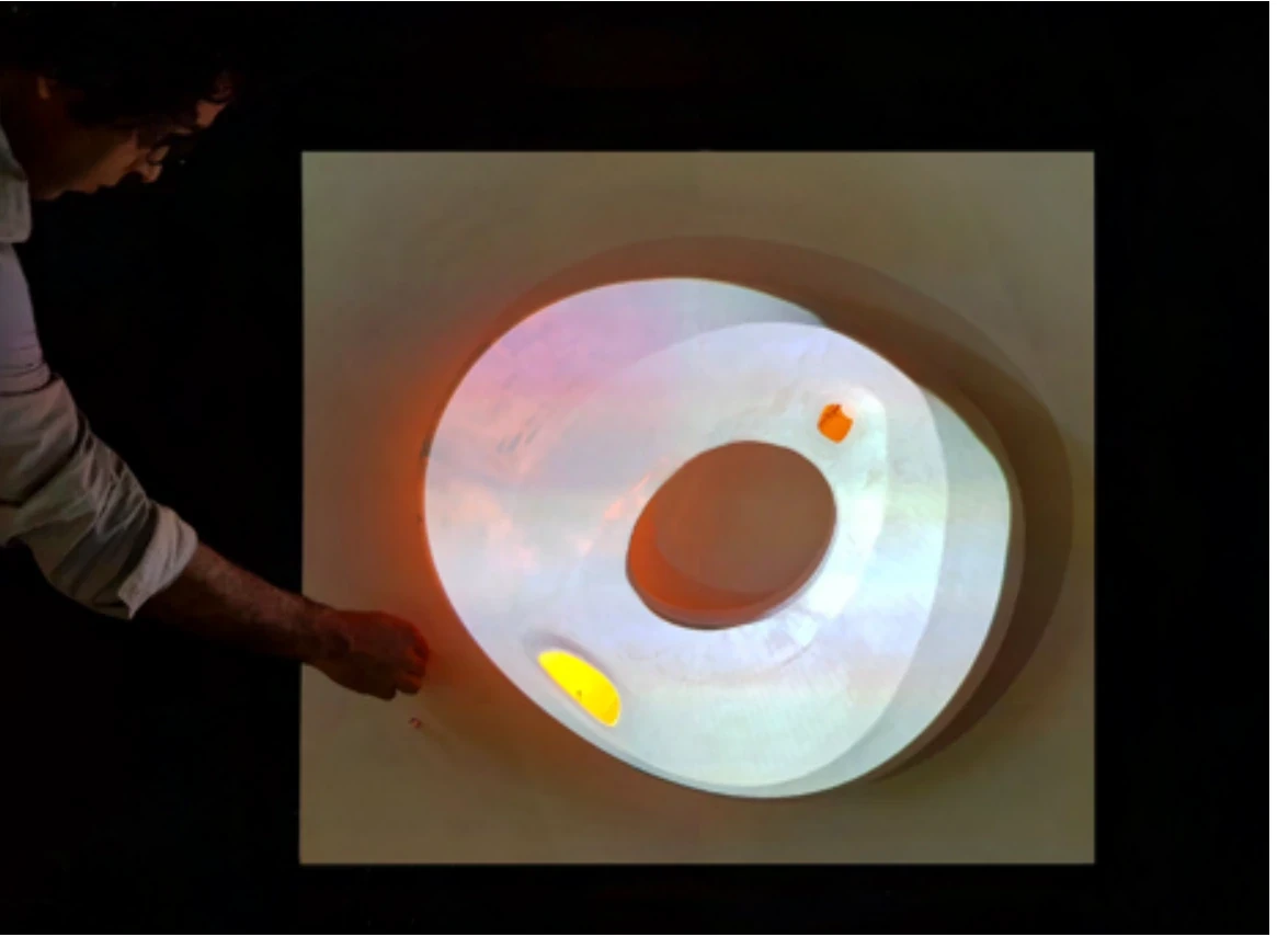 The torus lit from inside on a dark table, a hand pointing at its glowing opening.