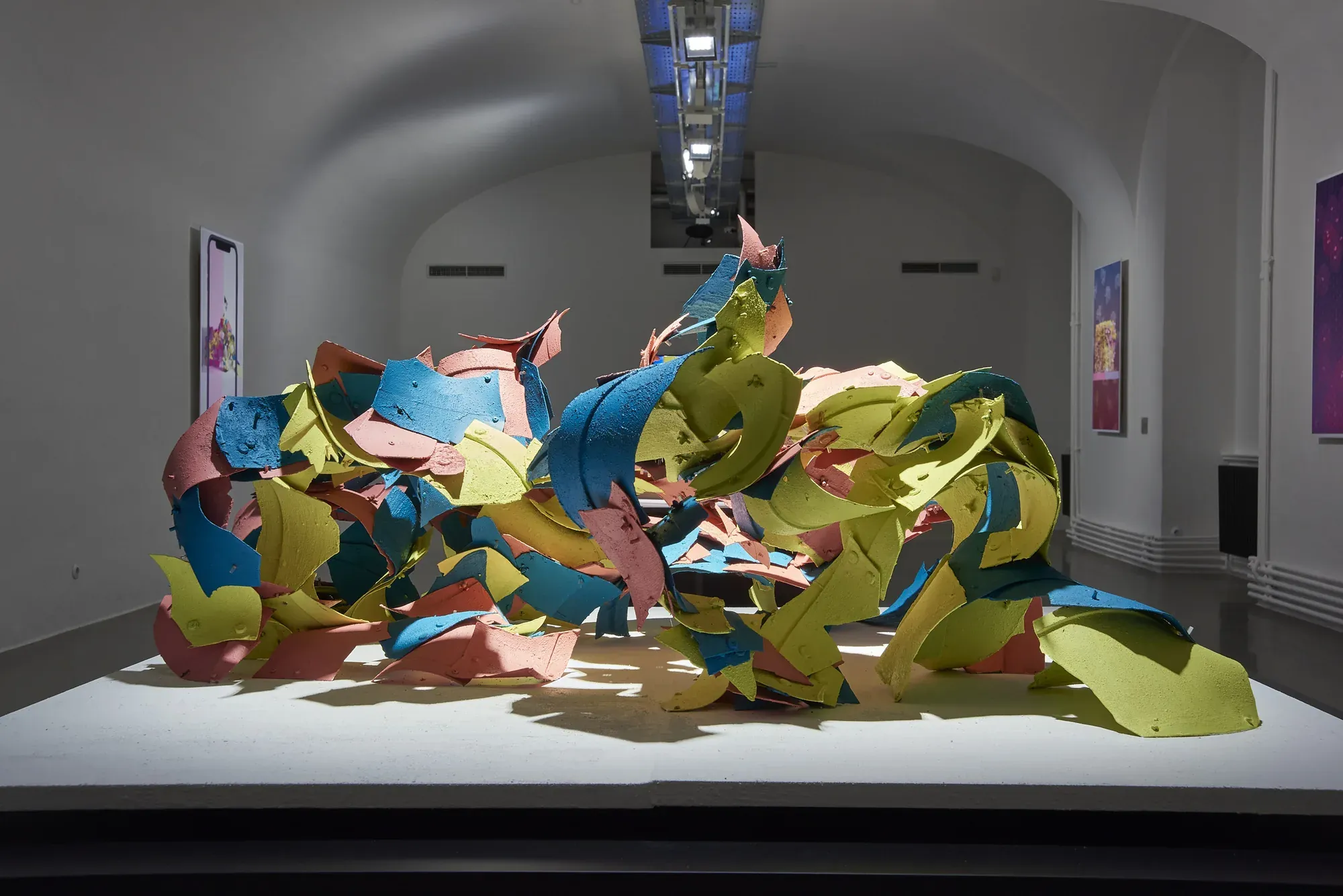 A large shell of colourful plastic fragments on a white plinth in a dim vaulted gallery, with framed posters on the walls.