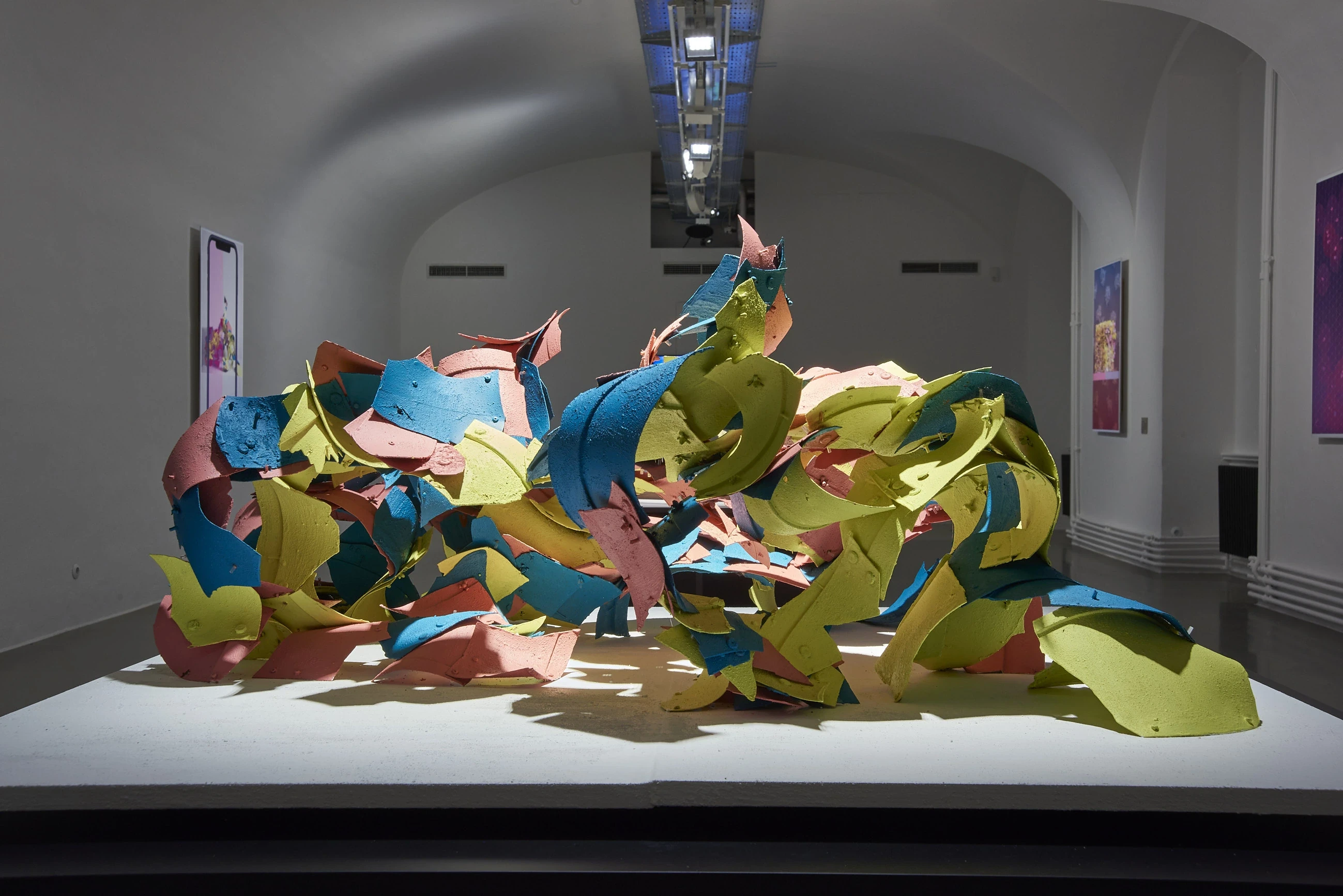 A large shell of colourful plastic fragments on a white plinth in a dim vaulted gallery, with framed posters on the walls.