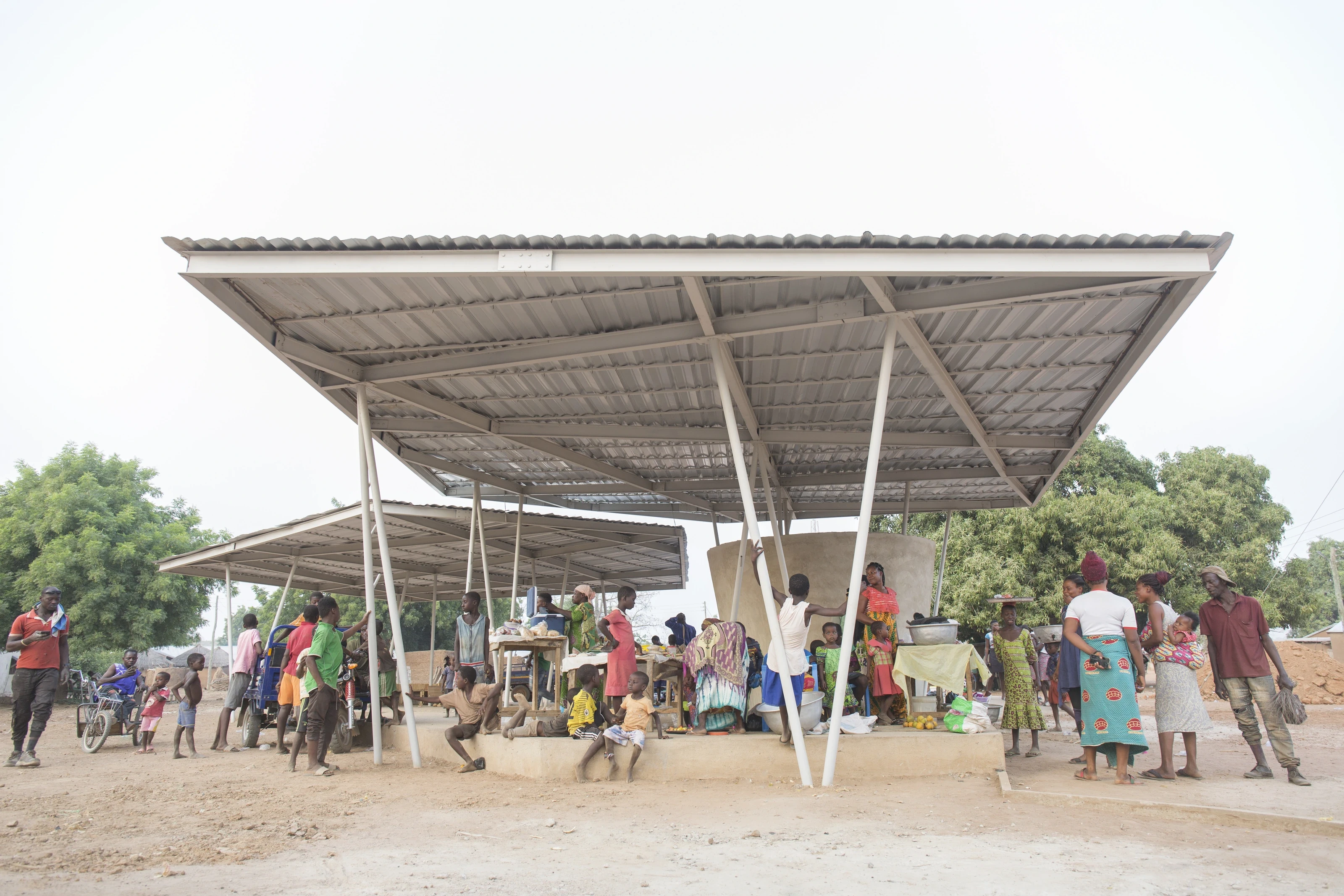 Traders and shoppers under the market's two wide, light steel roofs on slender white columns, with trees behind.