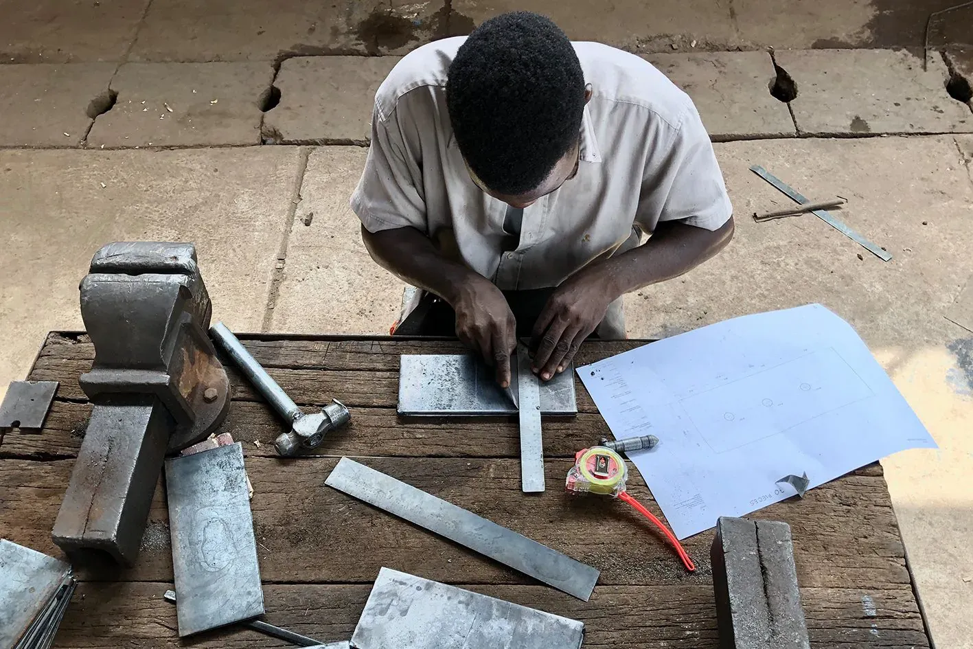 A worker at a bench cutting and marking steel plates, with a vice and tools.