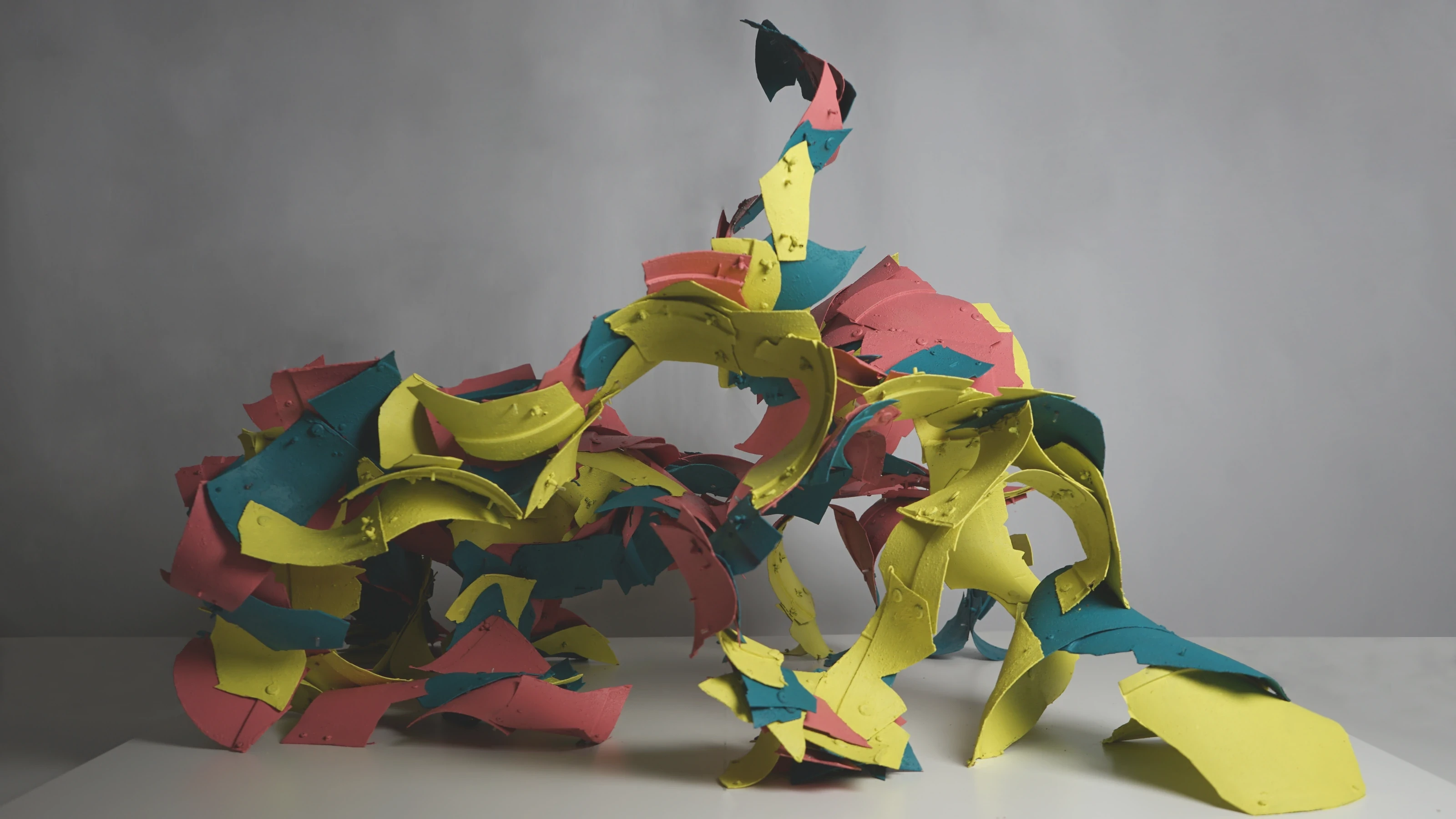 A tall, open shell built from hundreds of curved red, yellow and teal plastic fragments, photographed against a grey wall.