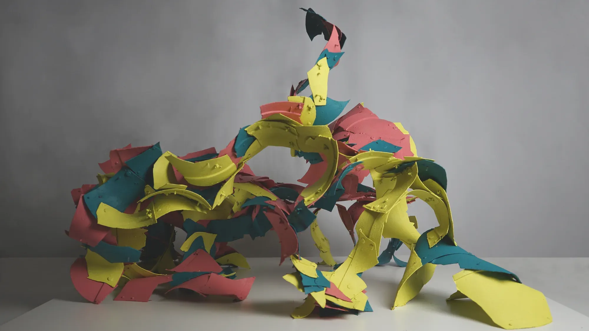 A tall, open shell built from hundreds of curved red, yellow and teal plastic fragments, photographed against a grey wall.