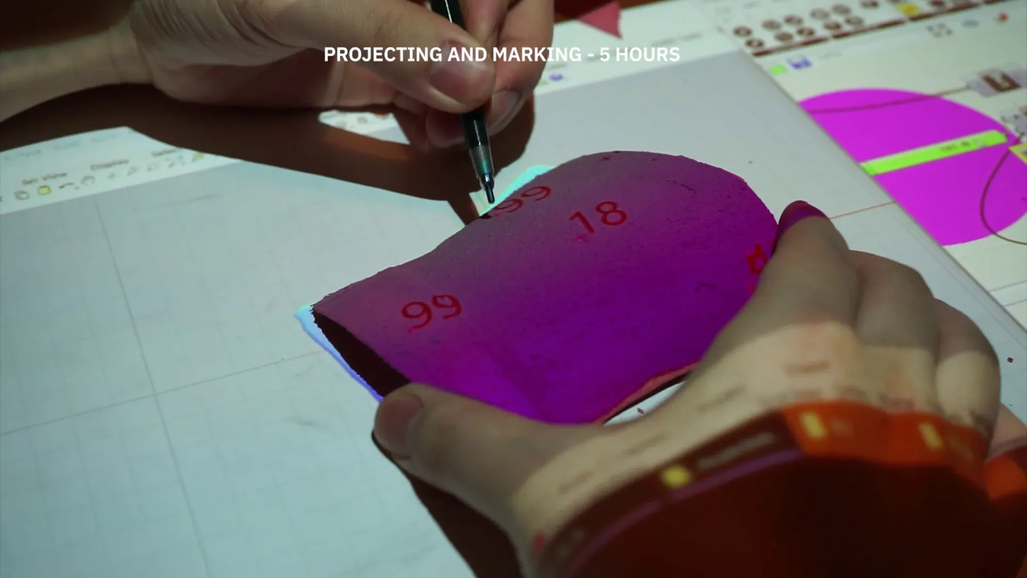 Close-up of two hands marking a pink plastic fragment with a pen; numbers 18 and 99 are projected onto it. Overlay text reads “Projecting and marking — 5 hours”.