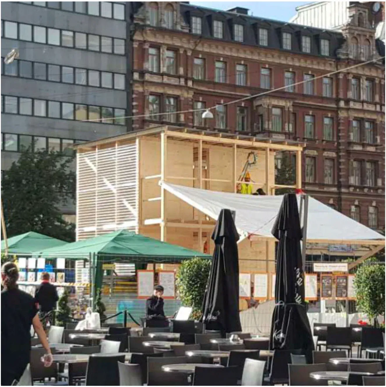 A timber shelter standing in a Helsinki square, café umbrellas and historic buildings around it.