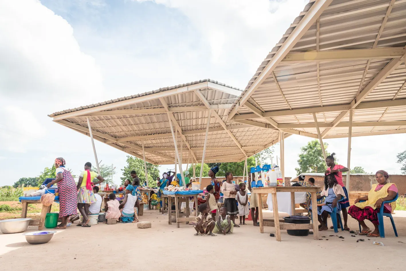 Traders and shoppers sitting under the market's wide, light steel roof on a bright day.