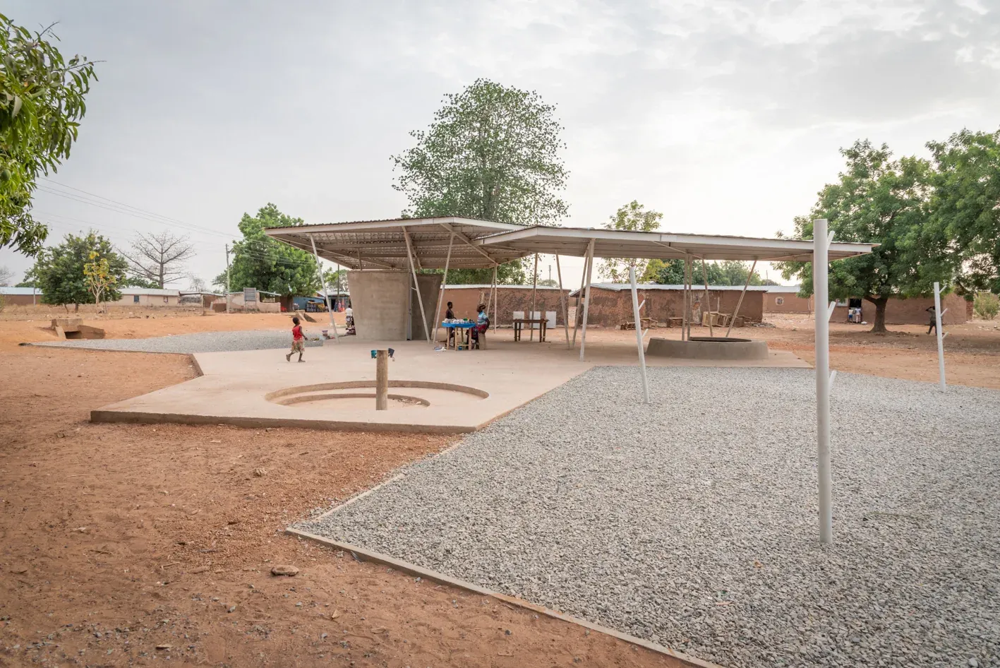 Wide view of the finished market: a light steel roof on slender columns over a gravel floor, with trees and low buildings behind.